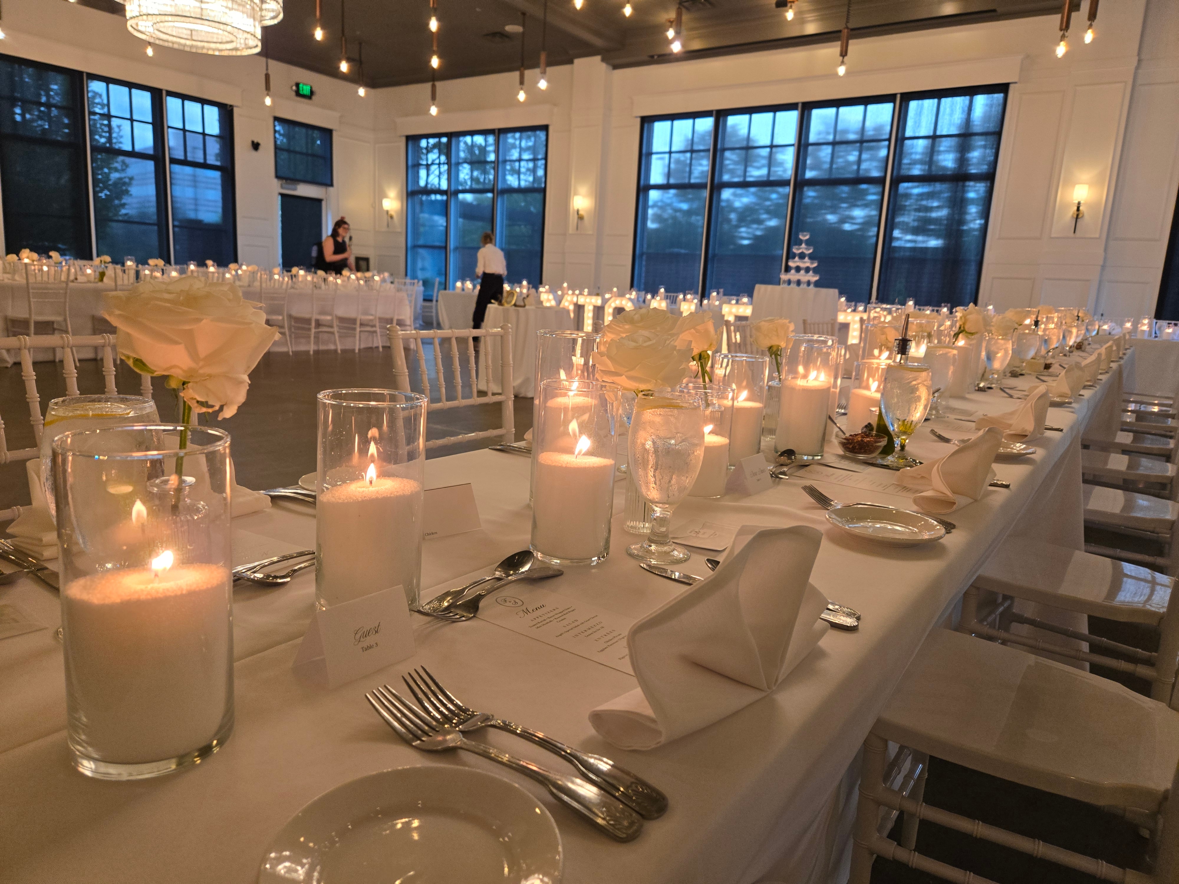 Long reception table with white linen, glass cylinder candlelight, and ivory roses