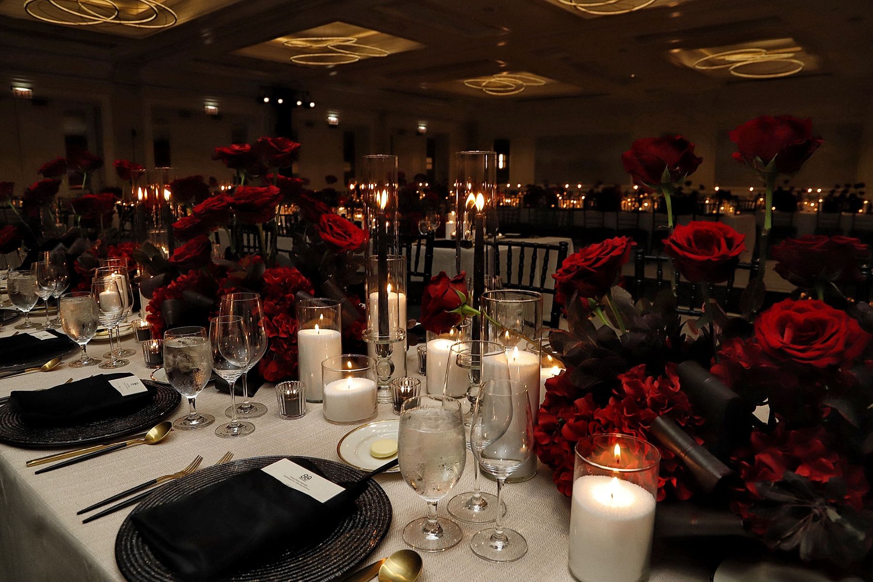 Pearled wax hurricanes and glass-enclosed tapers layered along a banquet table with red roses