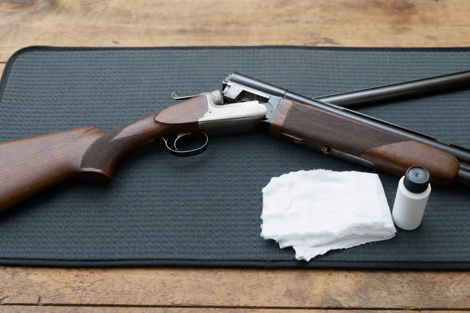 Unloaded break-action firearm on a protective mat with a cloth and unlabeled bottle