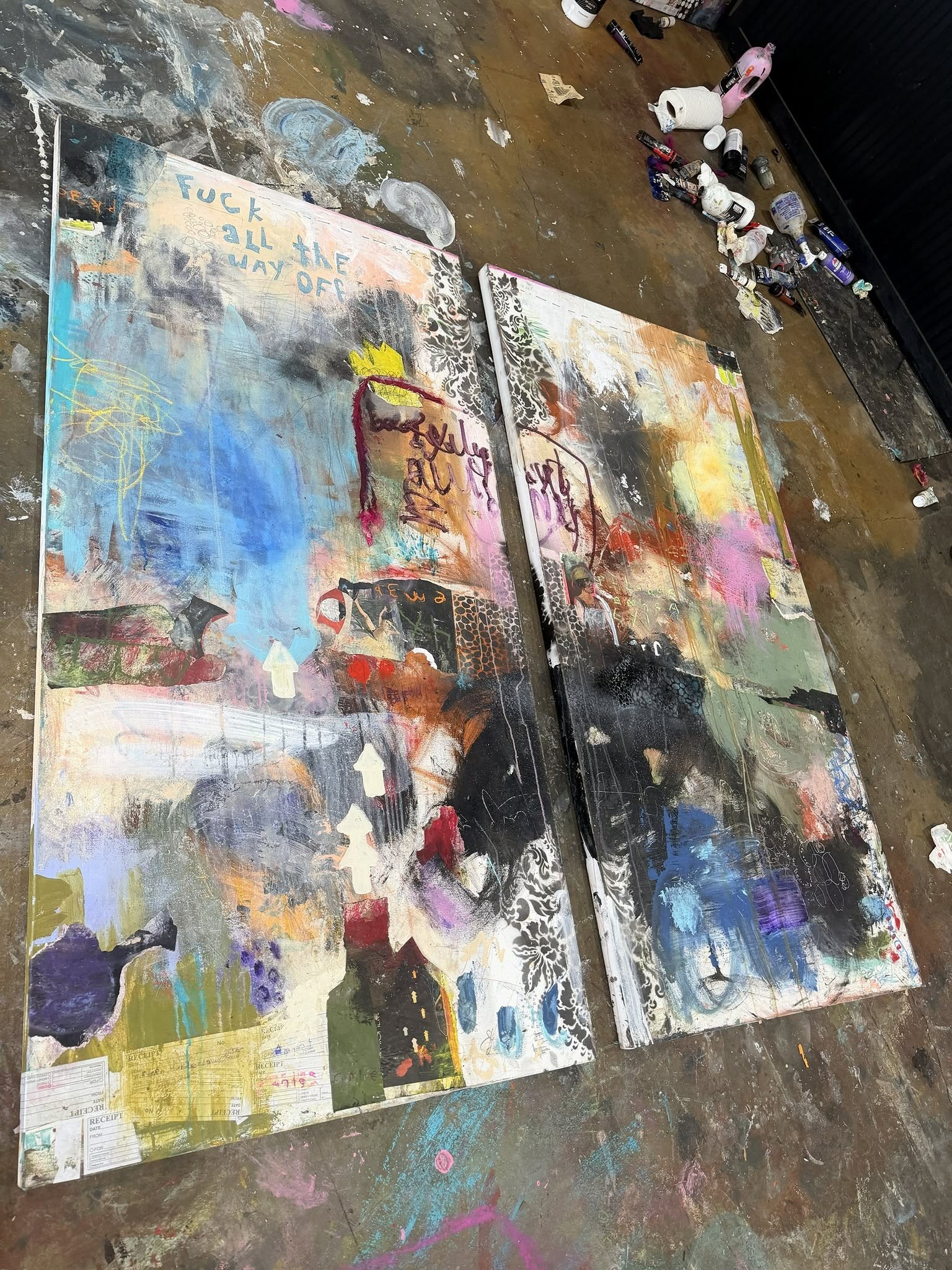 Two colorful mixed media canvas panels on Michel Keck’s paint-marked studio floor, with collage, handwritten text, blue, pink and yellow layers