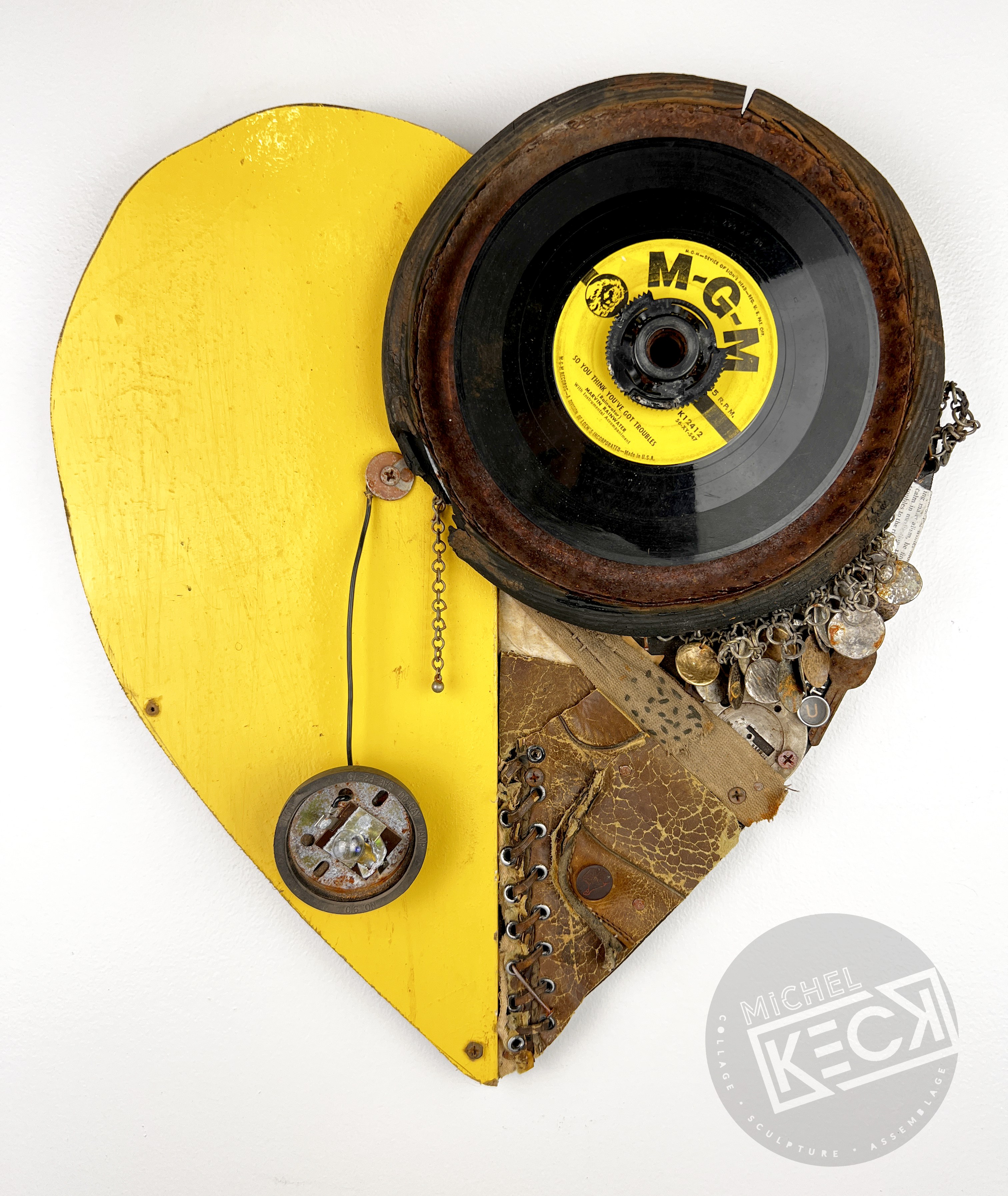 Mixed media heart-shaped wall sculpture by Michel Keck featuring a bright yellow painted wooden left half and a right half composed of a vintage record, worn leather, metal chains, and small found objects arranged in a textured collage.