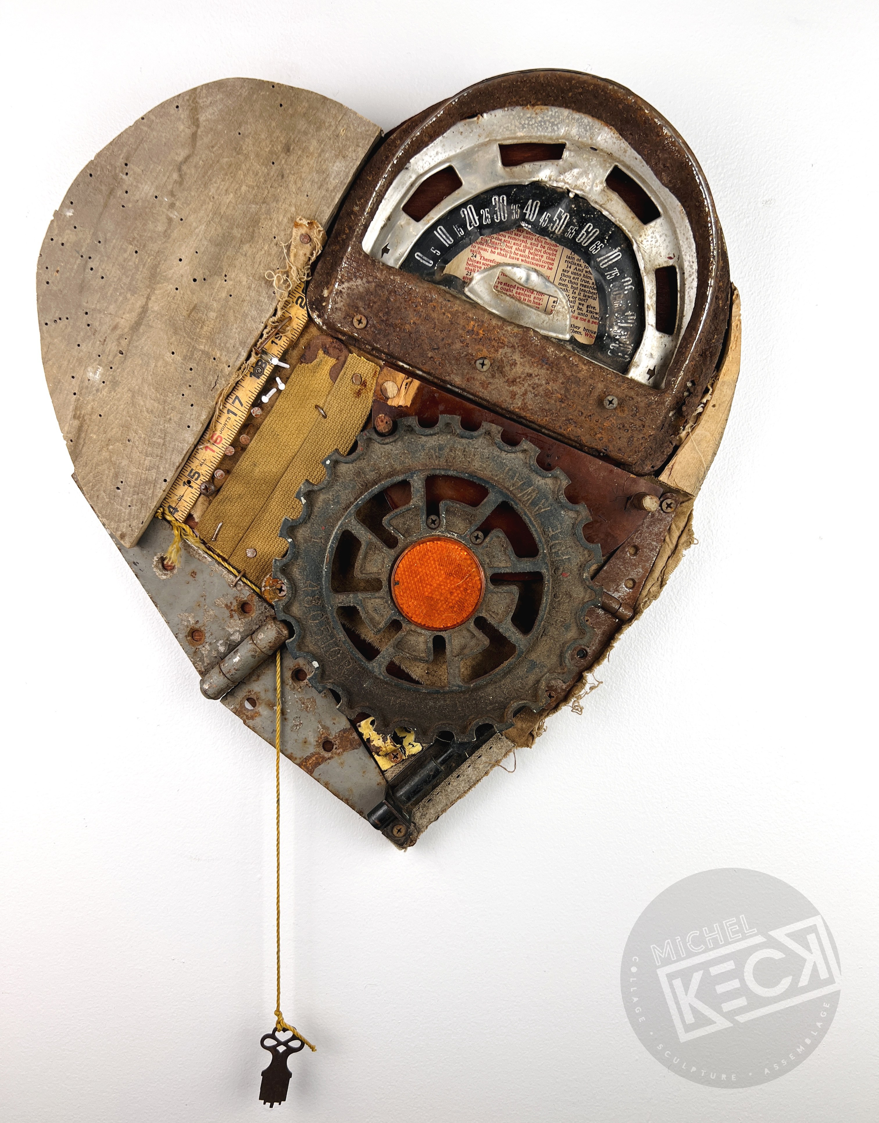 Heart-shaped assemblage by Michel Keck made from rusted metal, wood, and mechanical parts including a gear, a vintage gauge, and a dangling key on a chain, with a textured, weathered surface against a white background.