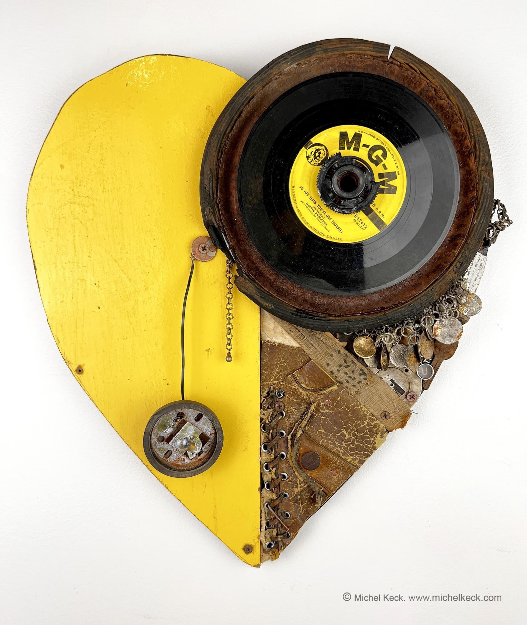 Mixed-media assemblage by Michel Keck shaped like a heart, featuring a bright yellow painted wooden left half and a right half composed of a vintage vinyl record, worn leather, metal chains, and small circular objects.