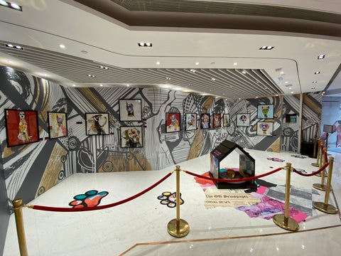 Michel Keck's dog art exhibit features colorful, stylized dog portraits framed and hung on a large wall with black and white geometric and organic patterns. The floor has paw prints and a small house sculpture surrounded by red ropes.