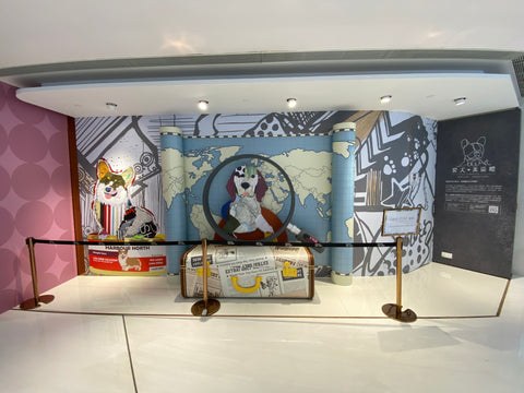 Michel Keck's dog art collage installation featuring a large circular frame with a stylized dog portrait, surrounded by patterned panels in muted blues, grays, and browns, with a sculpted dog figure and a suitcase prop in a gallery setting.