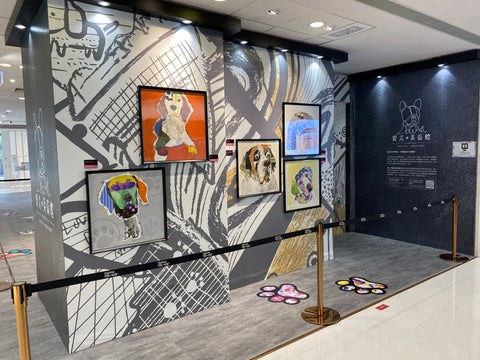Gallery installation featuring Michel Keck's dog-themed mixed-media collages mounted on a large black and white graphic wall. The artworks display colorful, stylized dog portraits in varied frames, with paw prints on the floor and a dark informational panel nearby.