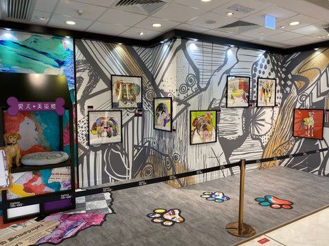 Michel Keck dog art show featuring colorful dog portraits in black frames mounted on a large wall mural with bold black and white abstract patterns. The floor has paw print designs, and a small dog bed with a stuffed dog is displayed nearby.