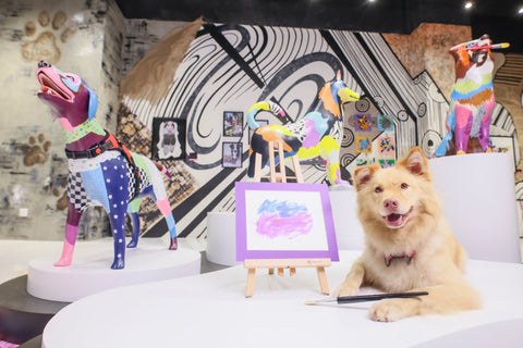 Colorful dog sculptures made from patchwork materials displayed on white pedestals in a gallery with black and white abstract wall art. A light brown dog lies on a table in the foreground holding a brush, surrounded by Michel Keck's dog collage art.