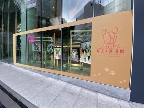 Michel Keck's dog collage art exhibit displayed behind large glass windows with colorful dog portraits on panels inside a modern building, featuring a beige frame with pink dog line drawings and paw prints on the right side.