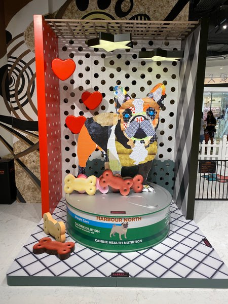 Three-dimensional dog collage by Michel Keck featuring a colorful patchwork dog figure with blue eyes, surrounded by oversized red hearts and bone shapes, set against a polka dot and grid patterned backdrop in a gallery space.