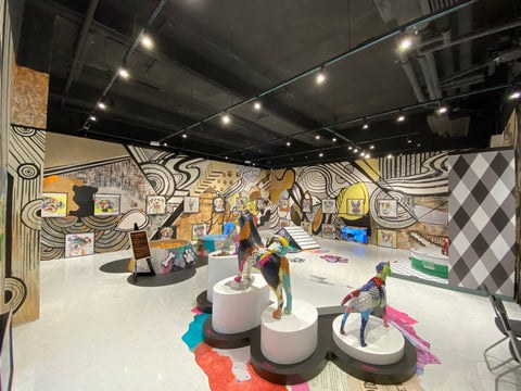 Interior view of a vibrant dog art exhibit by Michel Keck featuring colorful dog sculptures on white circular pedestals, surrounded by a large wall mural with bold black and white swirls, yellow accents, and various dog illustrations.