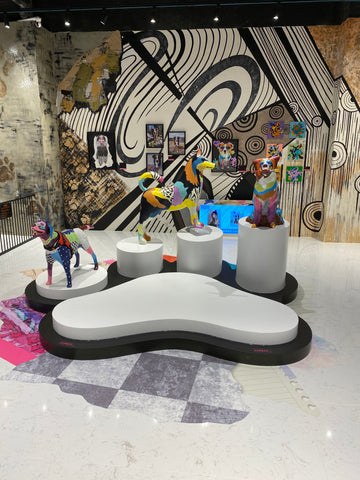 Colorful dog sculptures with vibrant patchwork patterns displayed on white cylindrical pedestals and a paw-shaped platform in a gallery space featuring a large black and white abstract mural with swirling lines and geometric shapes by Michel Keck.