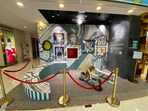 Michel Keck's dog art show installation features a curved, striped platform and a small dog sculpture on a skateboard, surrounded by red ropes and gold stanchions. The backdrop displays colorful framed dog-themed artworks against a large abstract mural.