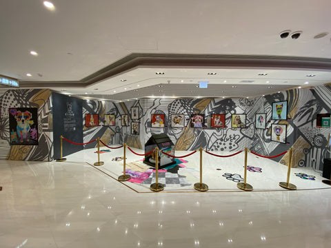 Michel Keck's dog art collage exhibit features colorful framed dog portraits arranged on a large wall with black and white abstract patterns. The floor has a painted doghouse and paw prints, all enclosed by gold stanchions with red ropes in a polished gallery space.