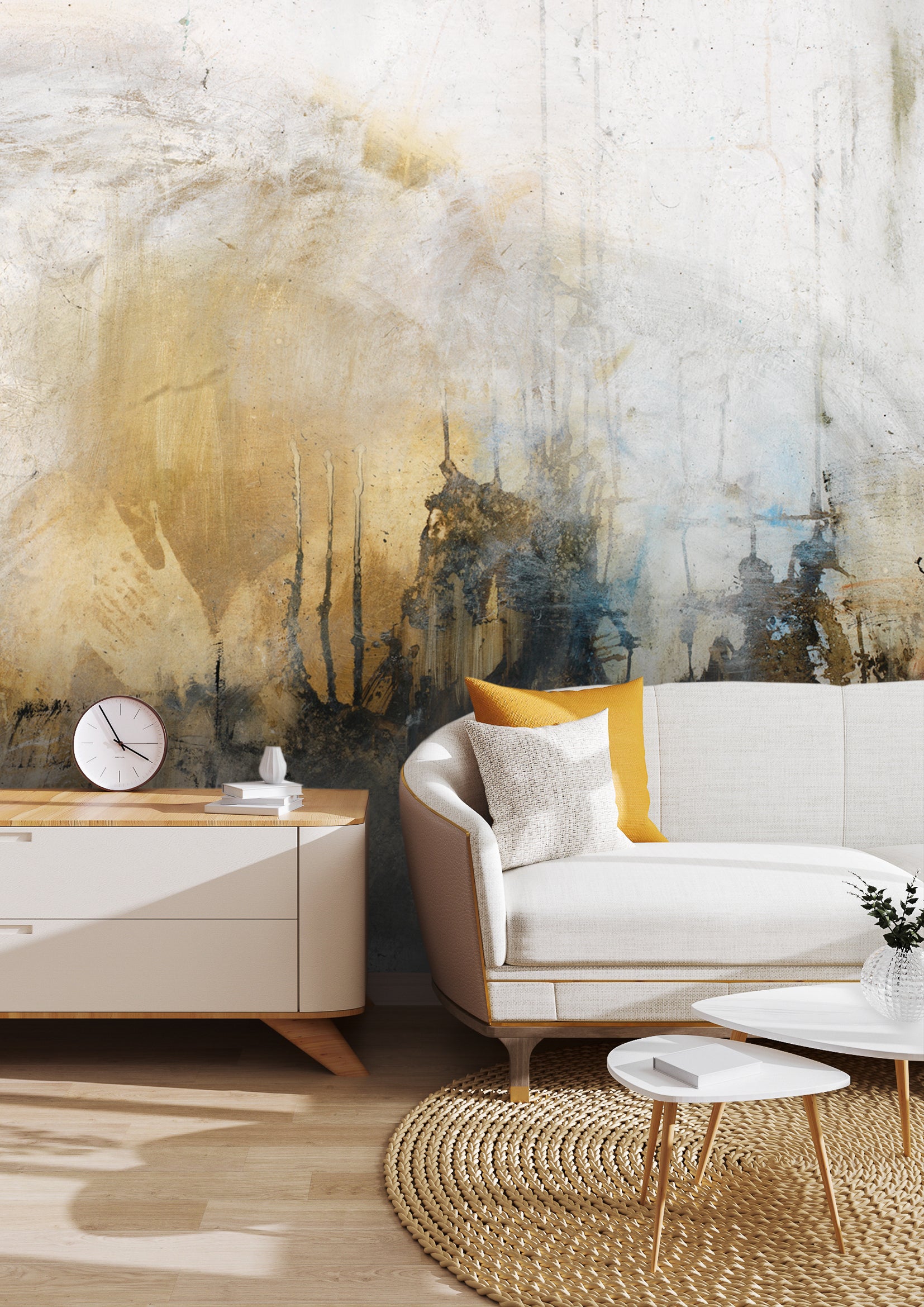 A modern living room featuring a large peel-and-stick wall mural by Michel Keck with abstract textures in muted gold, black, and blue tones. The space includes a white sofa with yellow and patterned pillows, a light wood sideboard, and round white coffee tables on a woven circular rug.