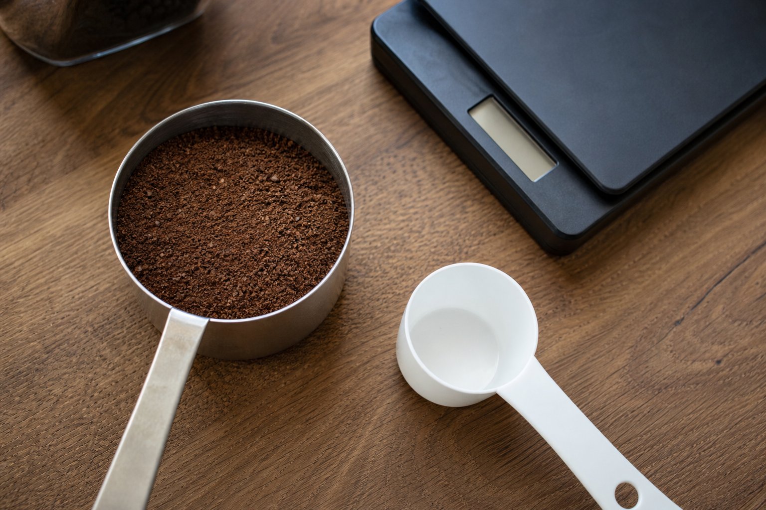 Ground coffee in a metal scoop beside a white measuring scoop and kitchen scale