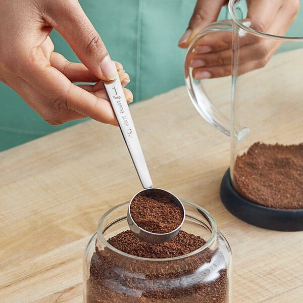 A level stainless steel coffee scoop filled with ground coffee beside a glass coffee jar