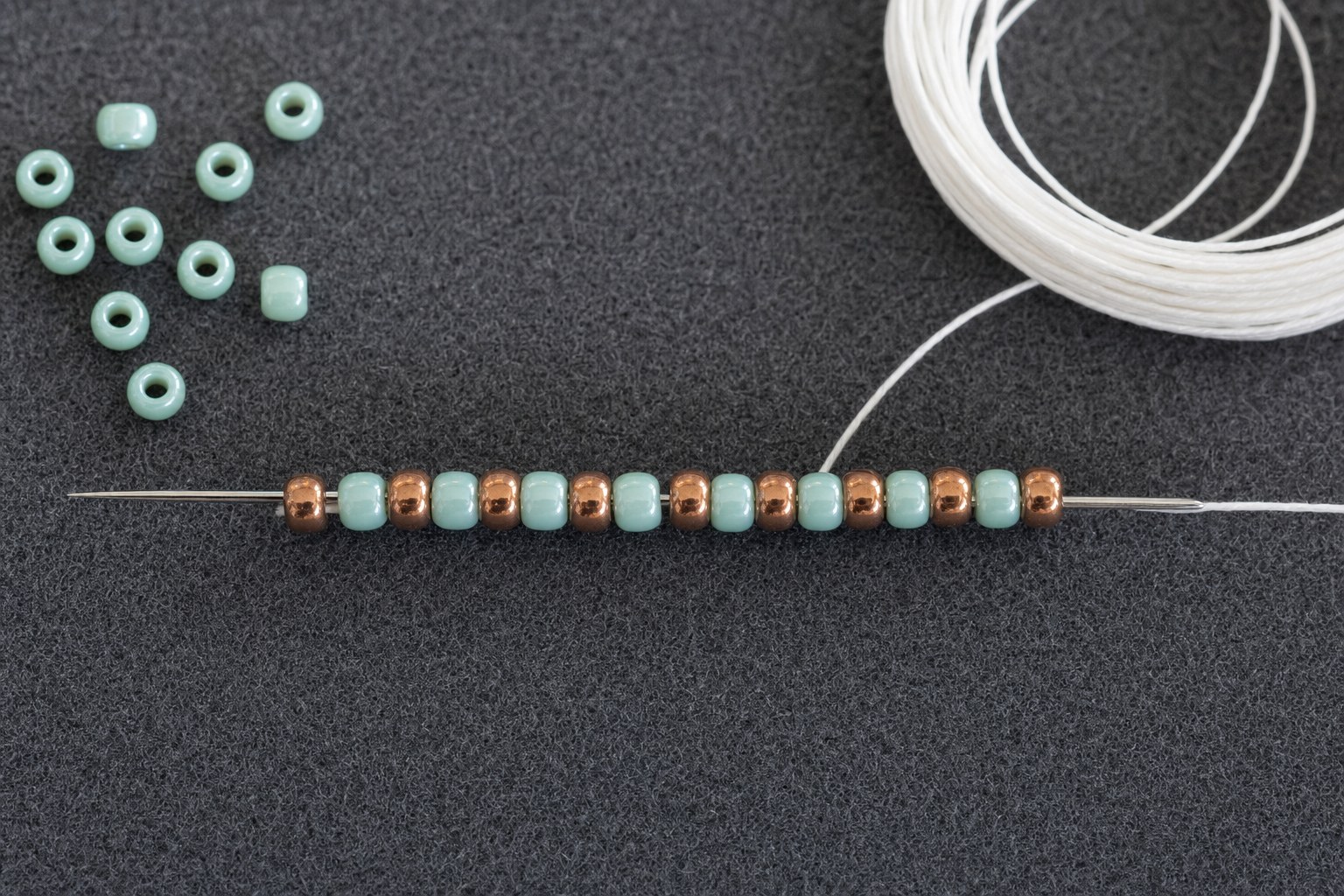A fine beading needle and white thread passing through small turquoise and copper seed beads