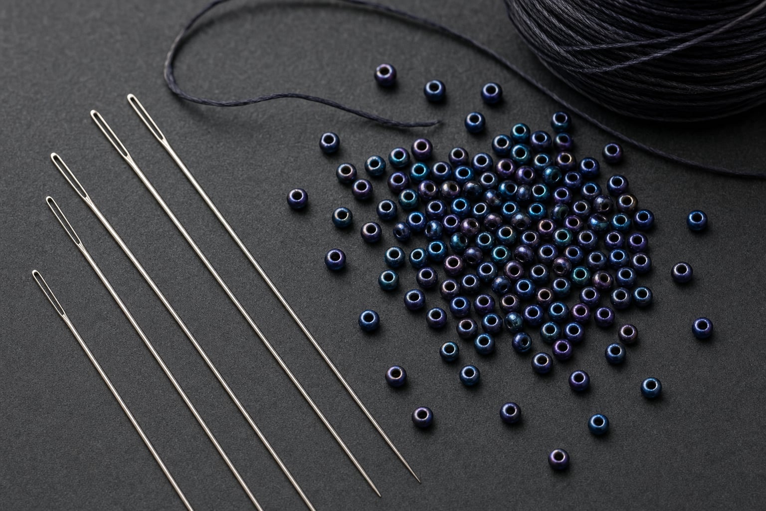 Fine beading needles beside dark thread and blue glass seed beads