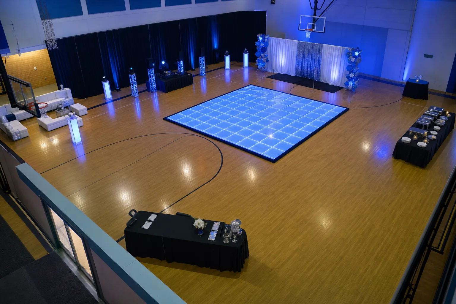 High-angle view of a school gym arranged with a central LED dance floor, seating, food tables, and a photo backdrop