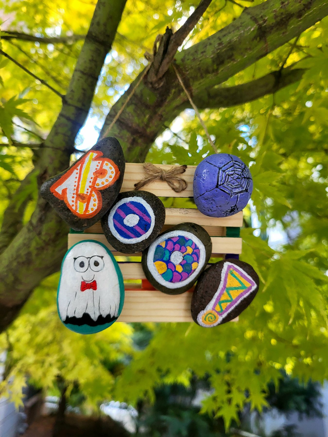 Six Halloween painted rocks spelling Boo! with a ghost and spiderweb, mounted on a wooden hanging backing outdoors