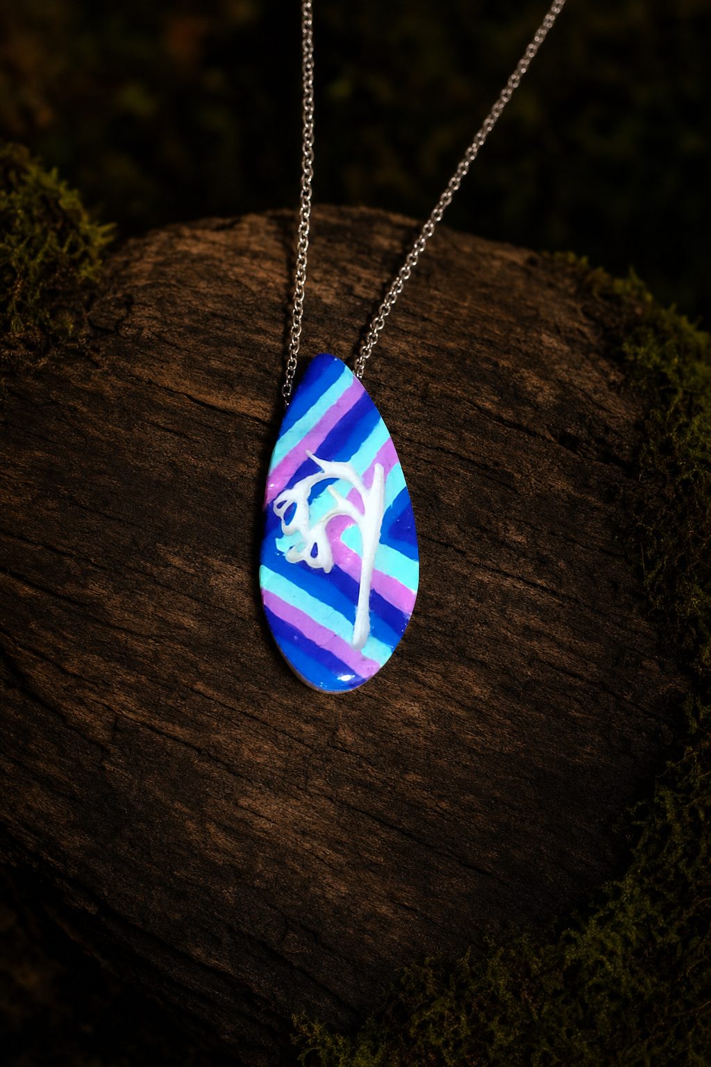 Striped teardrop clay pendant with raised white botanical detail on dark wood and moss
