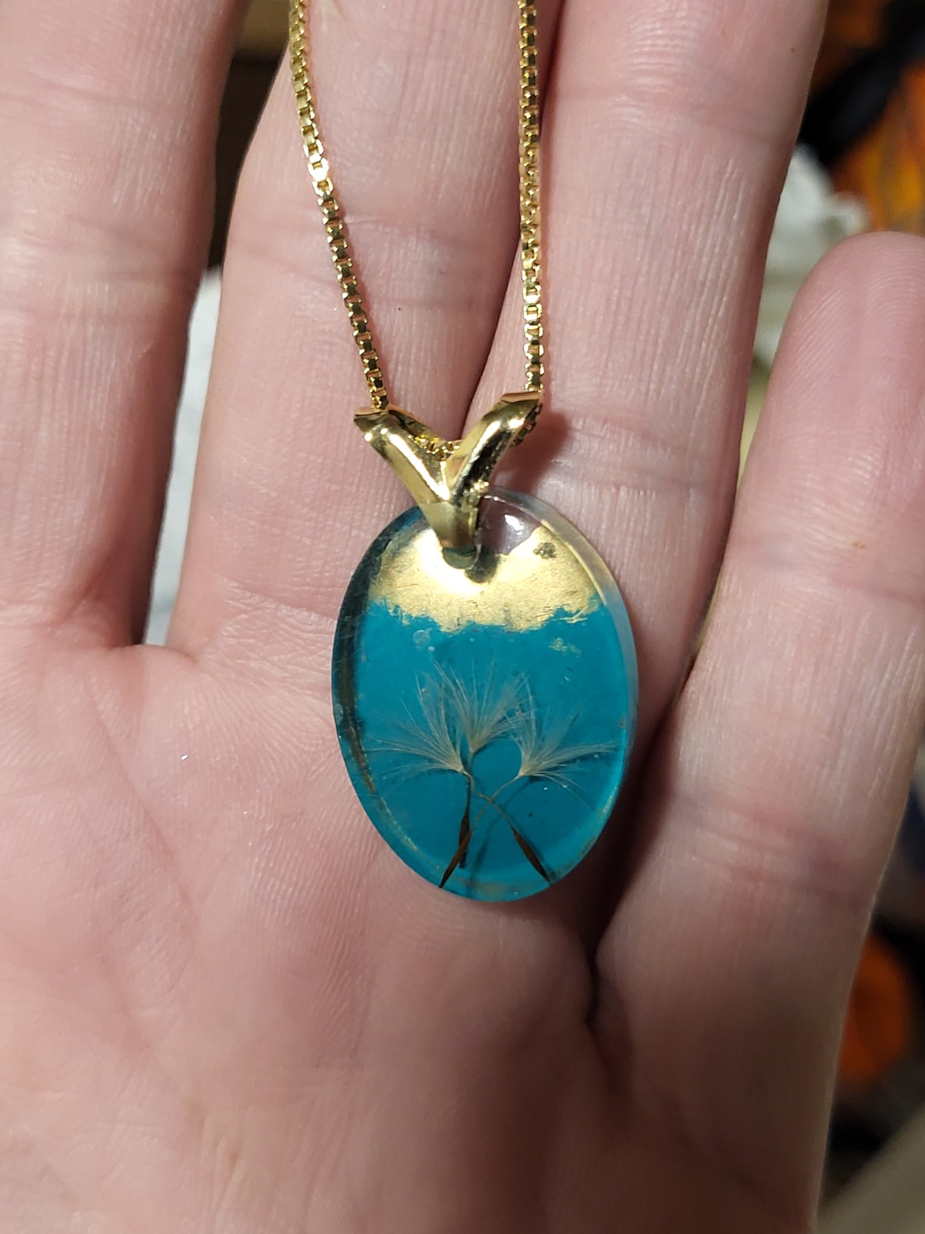 Teal oval dandelion wish pendant with gold-colored detailing, shown in a hand