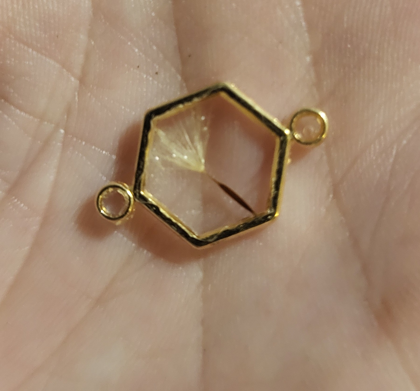 Small clear resin wish charm with a dandelion in a gold hexagon setting, shown in a hand