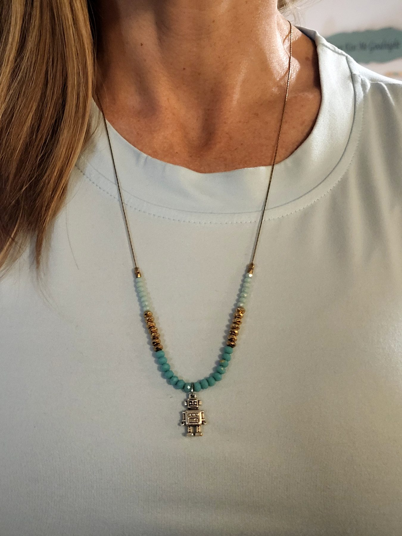 Robot pendant necklace with teal and gold beads, shown worn on a light shirt