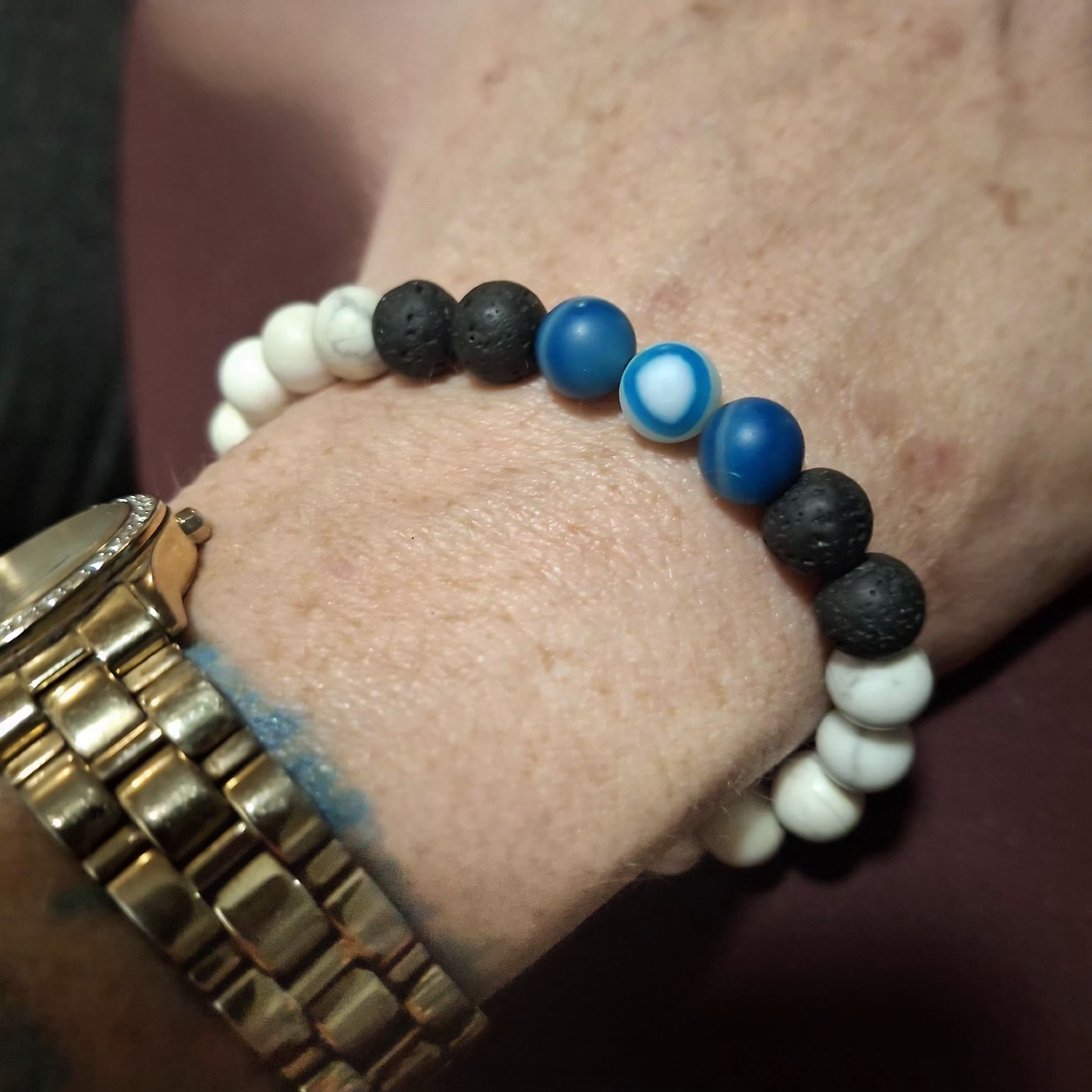 Unisex evil eye bracelet worn on a wrist, with white howlite, black lava, and banded blue agate beads