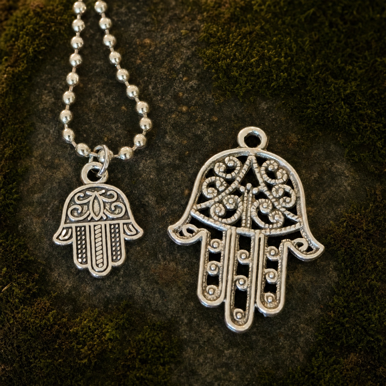 Smaller Hamsa hand charm on a simple ball chain beside a larger openwork Hamsa pendant, displayed on dark stone and moss