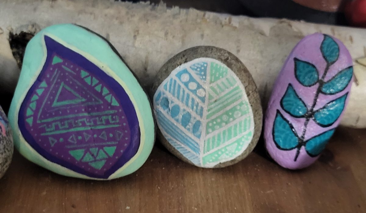 Three hand-painted stones with geometric leaf designs in purple, mint, blue, and teal, displayed on a wooden shelf