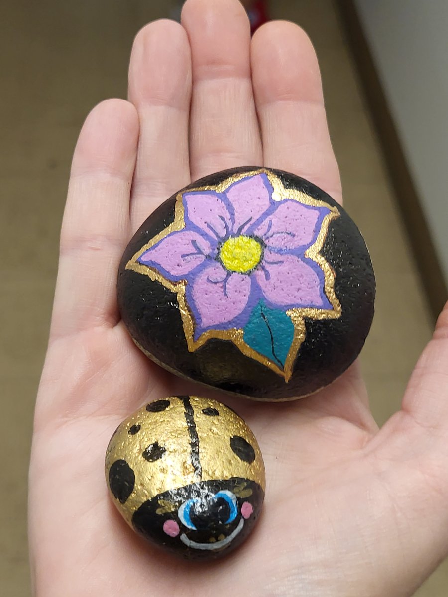 Metallic-gold ladybug and a purple flower-painted rock with gold edging, held in a hand to show scale