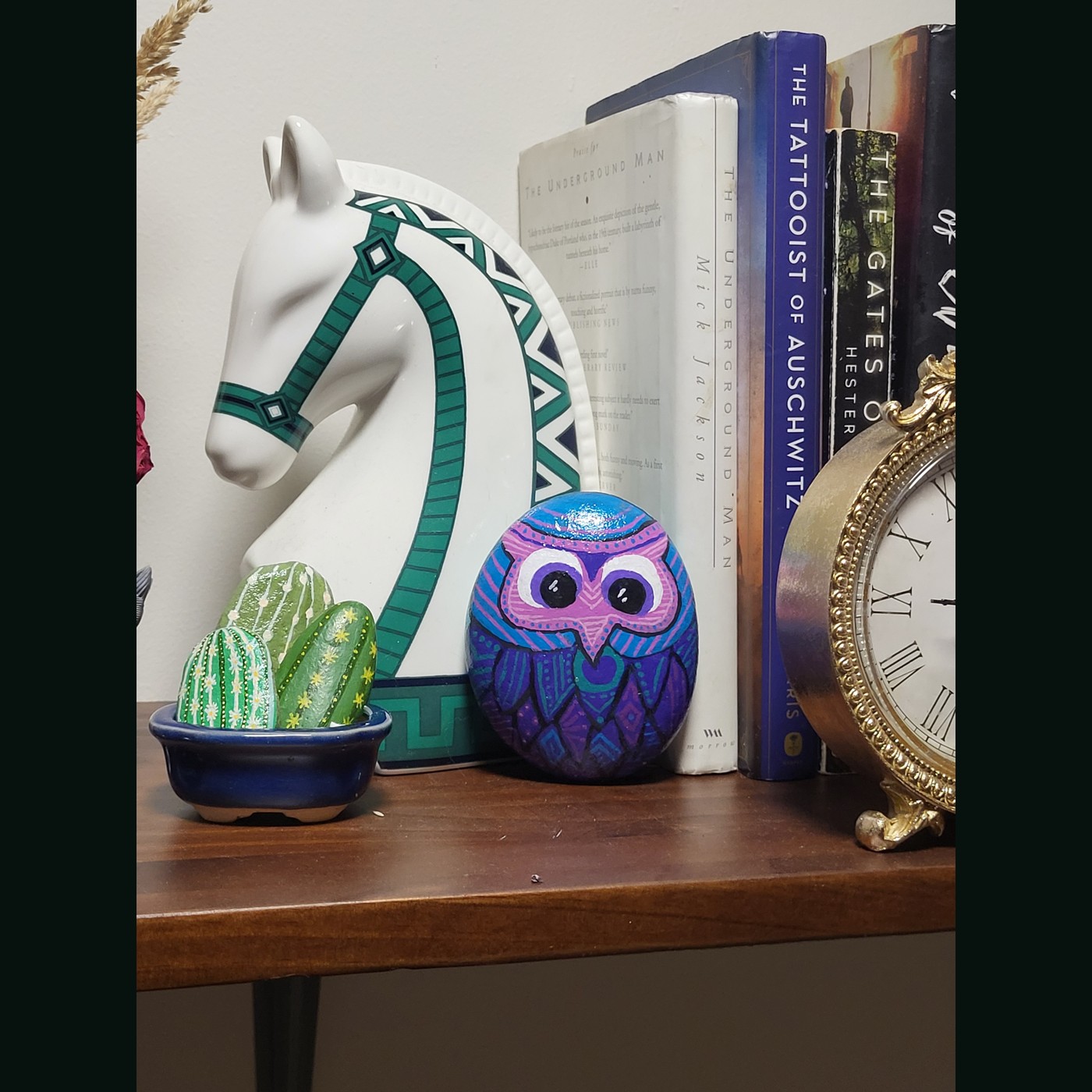 The same large painted owl rock displayed on a wooden shelf beside books, a decorative horse and a clock