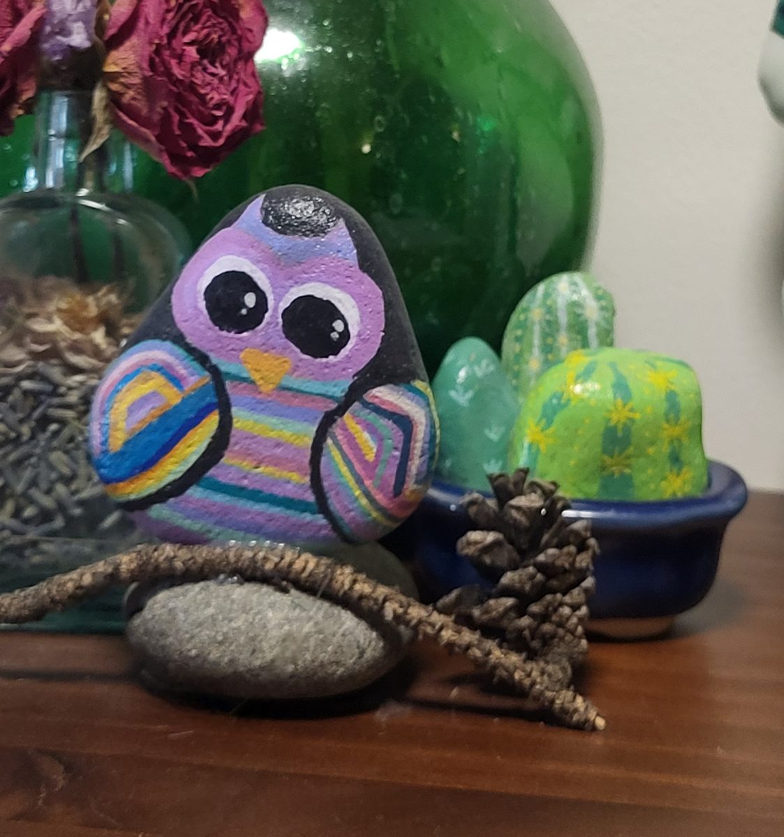 Colorful hand-painted owl stone mounted upright on a branch above a stone base, displayed on a wooden shelf