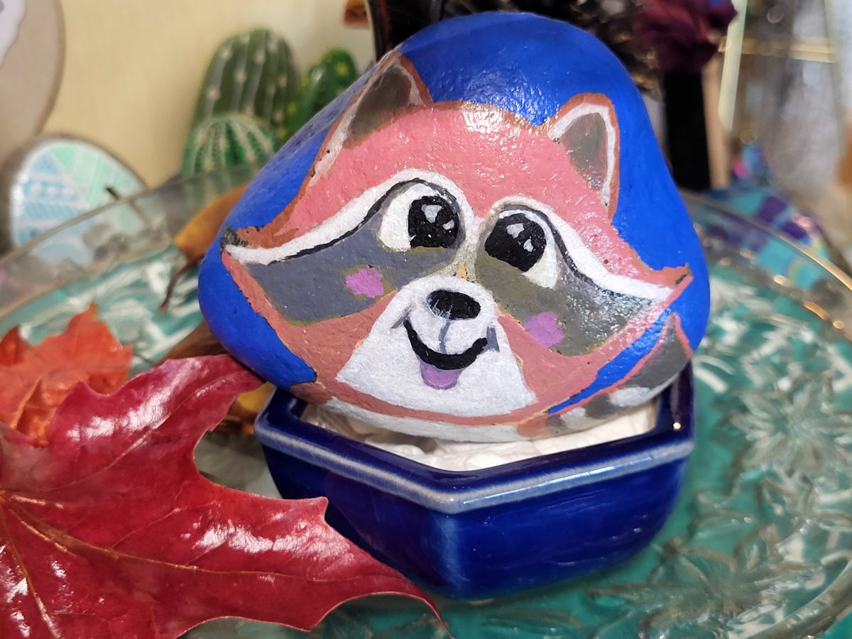 Smiling raccoon painted in pink, gray, and white on a blue stone