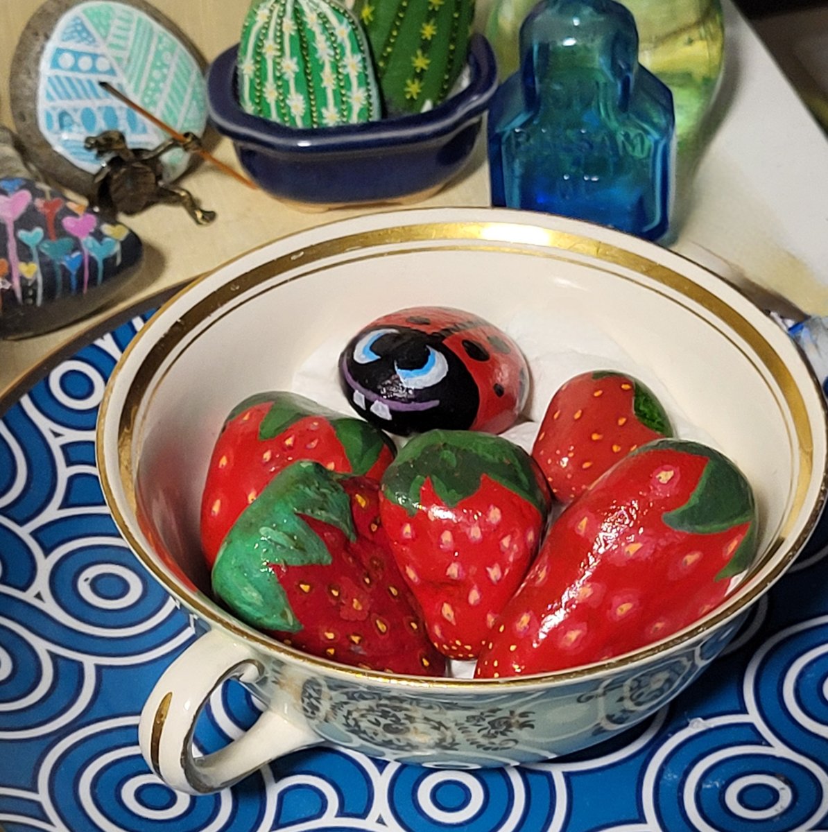 Five hand-painted strawberry stones and one smiling ladybug stone arranged in a decorative teacup