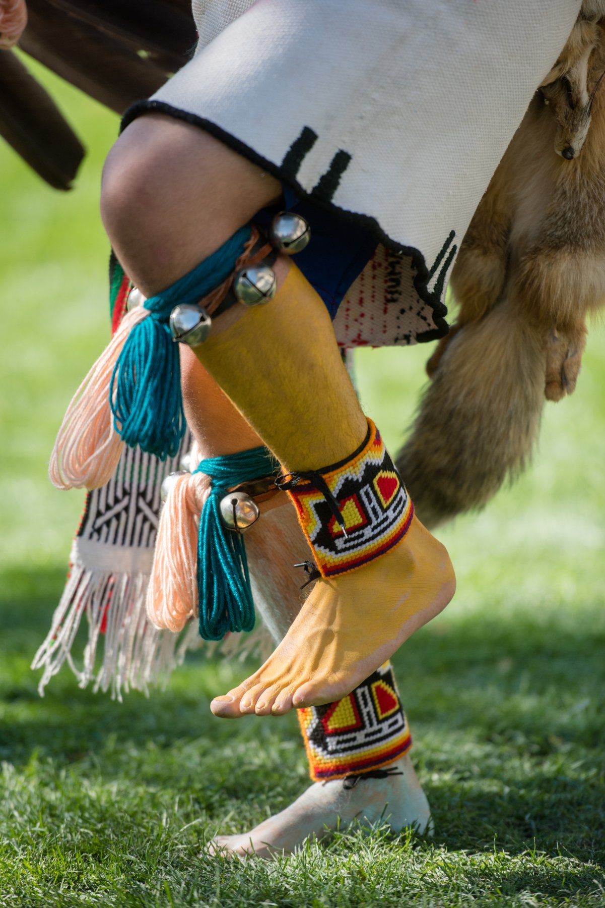 Close-up of a dancer’s feet, beaded leg bands, bells, and fringe in motion on grass