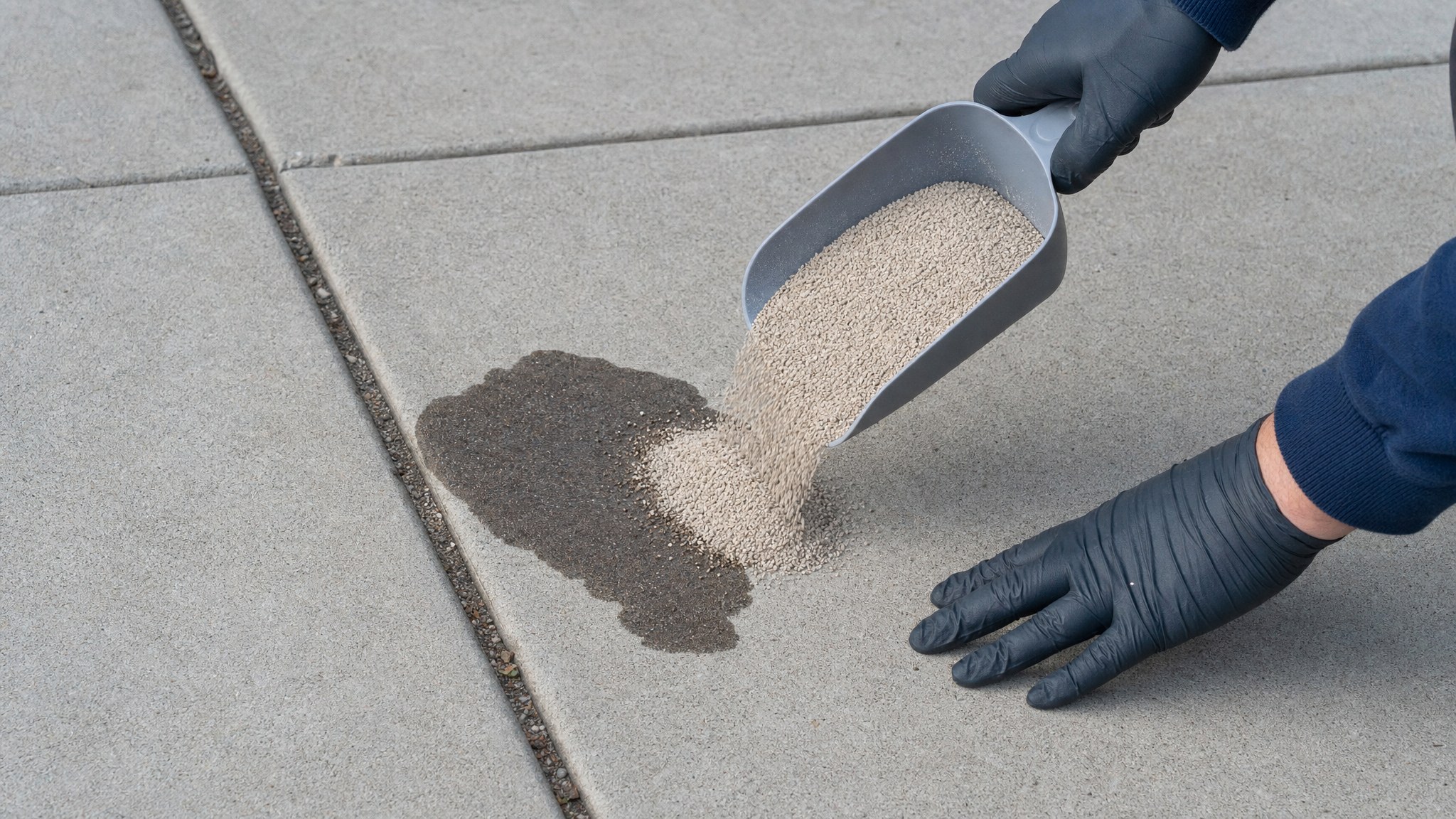 Gloved hands covering a fresh oil spot on a concrete driveway with absorbent material