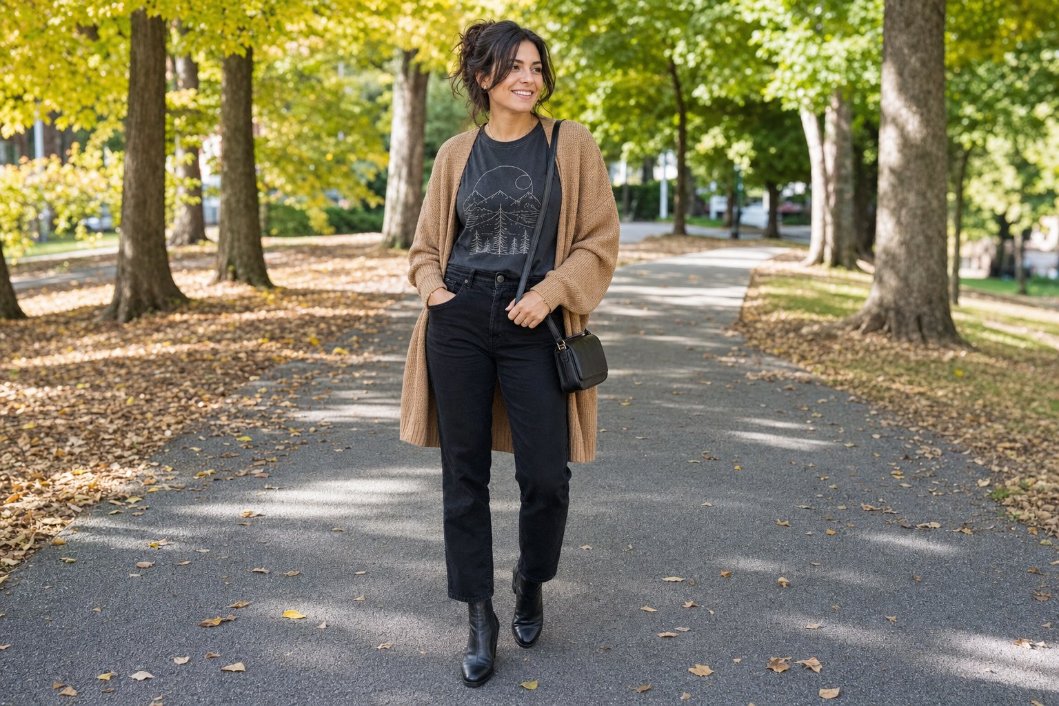Woman wearing a charcoal graphic T-shirt, black jeans, black ankle boots, and a camel cardigan on an early fall park path