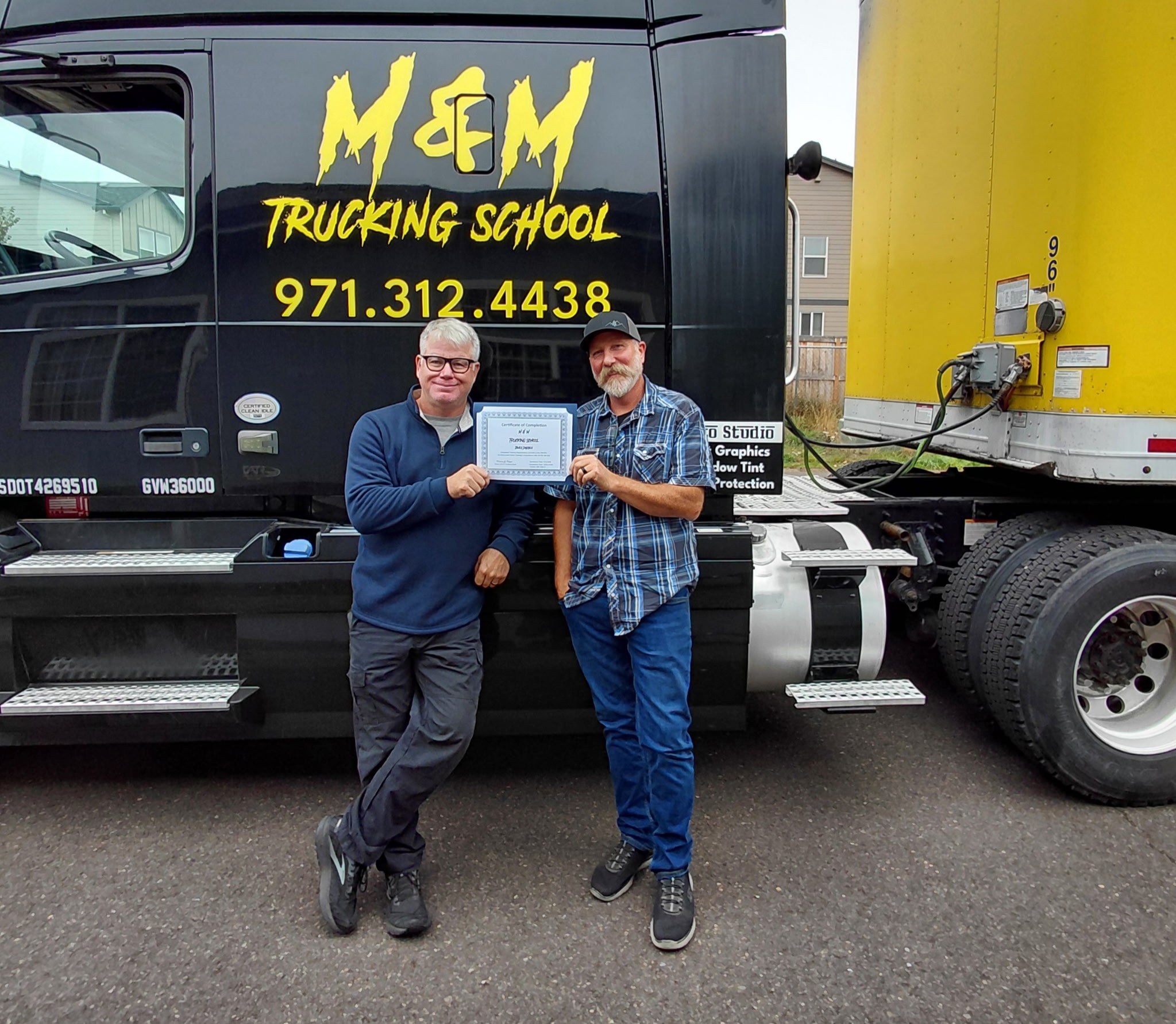 M&M Trucking School graduate holding his certificate with a companion beside a training truck