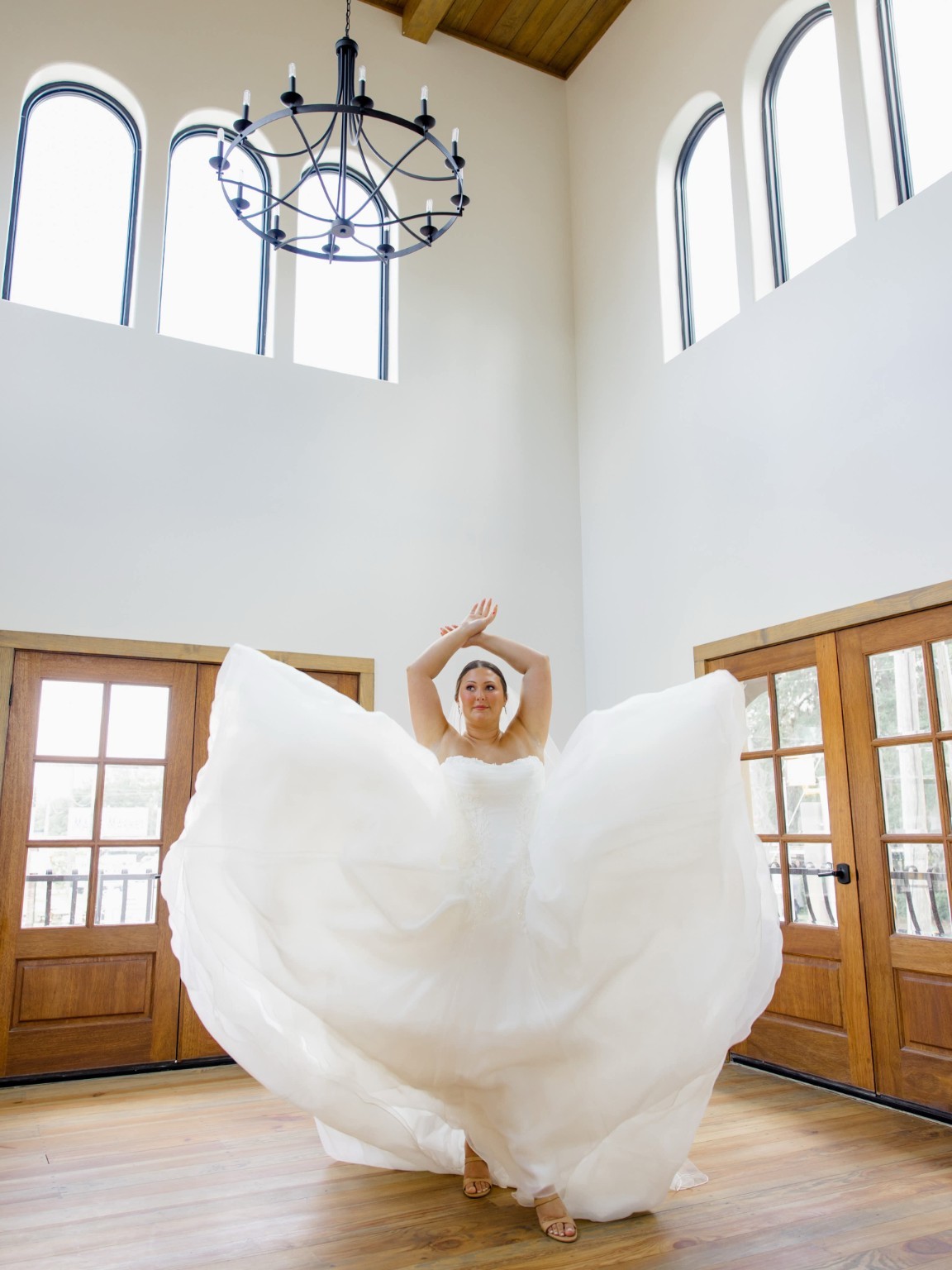 Bride with an elegant updo lifting her flowing wedding gown beneath tall arched windows