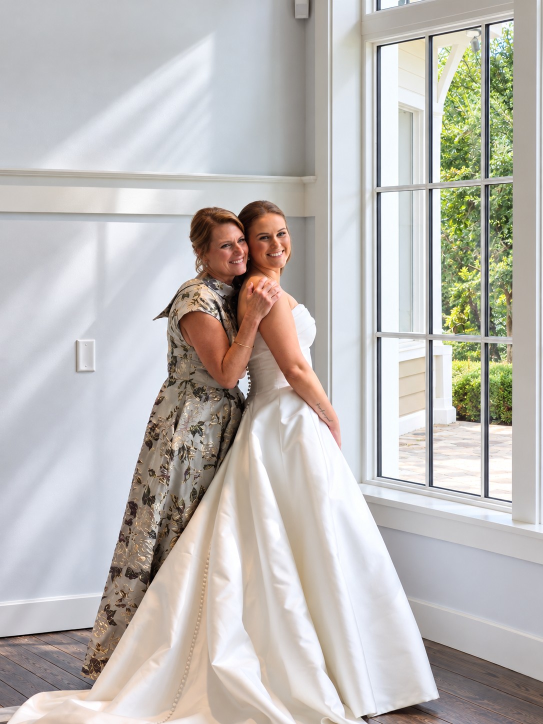 Bride and her mother embracing beside a sunlit window on her wedding morning