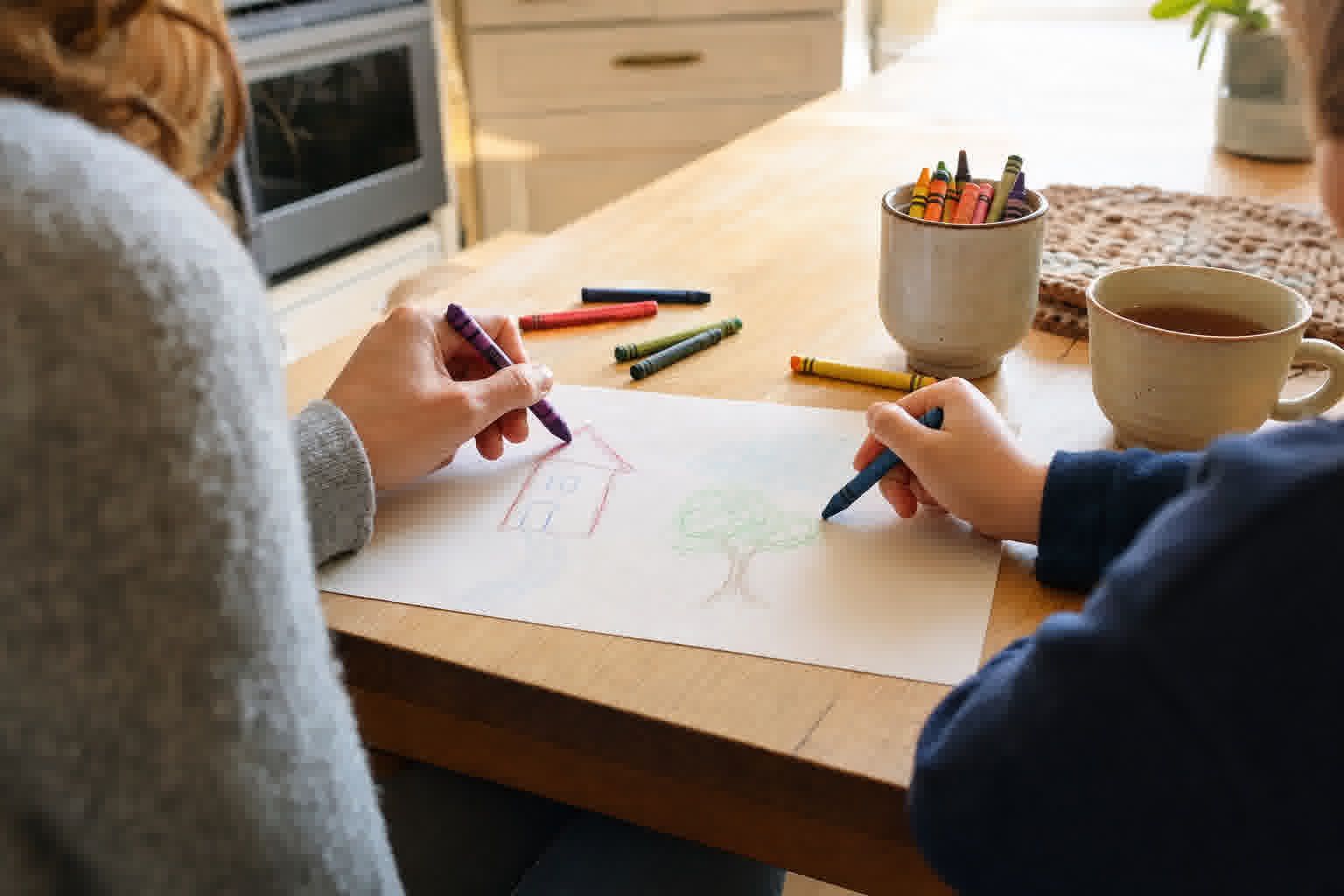 A parent and child drawing together at a kitchen table with crayons.