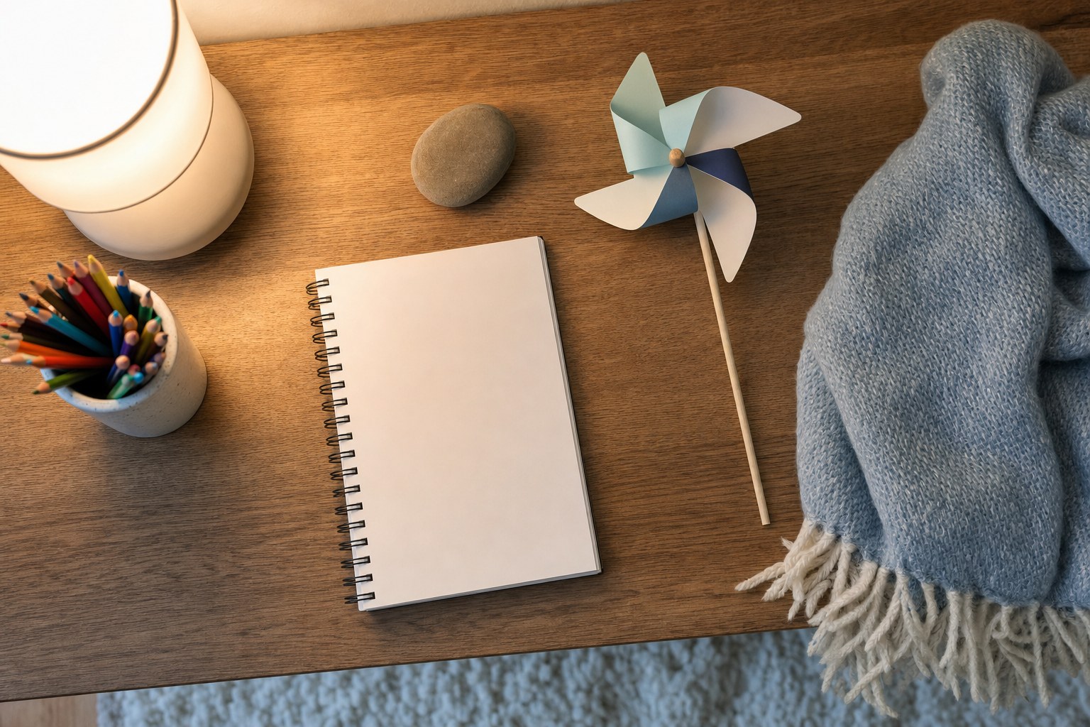 A blank notebook, colored pencils, smooth stone, paper pinwheel, lamp, and soft blanket arranged for a child's calming routine.