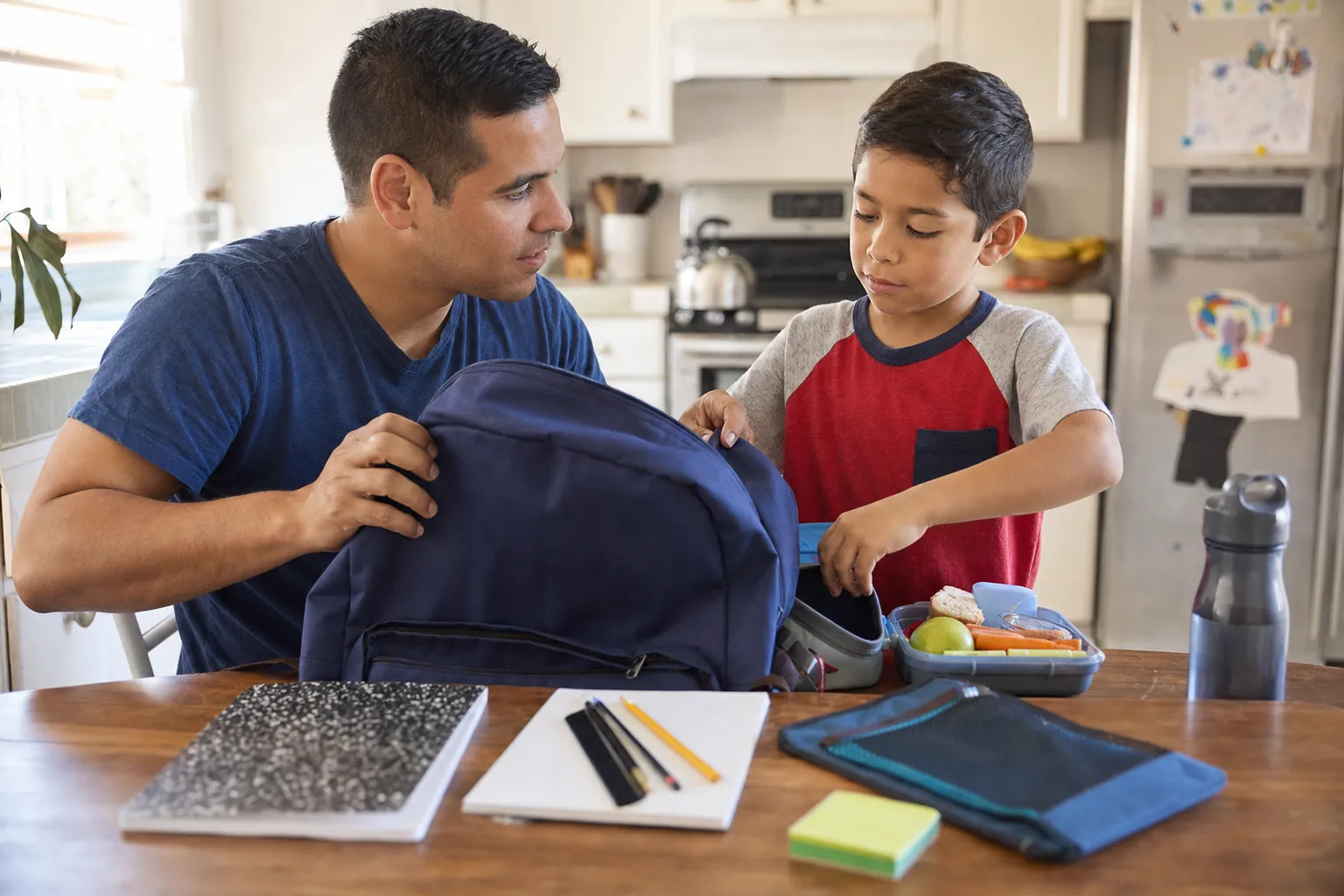 A parent and child pack a backpack together at the kitchen table before school.