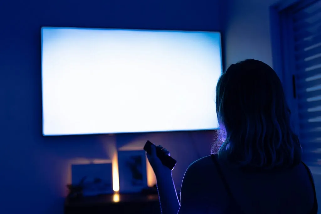 A woman sitting alone in a dark room, looking toward a glowing television.