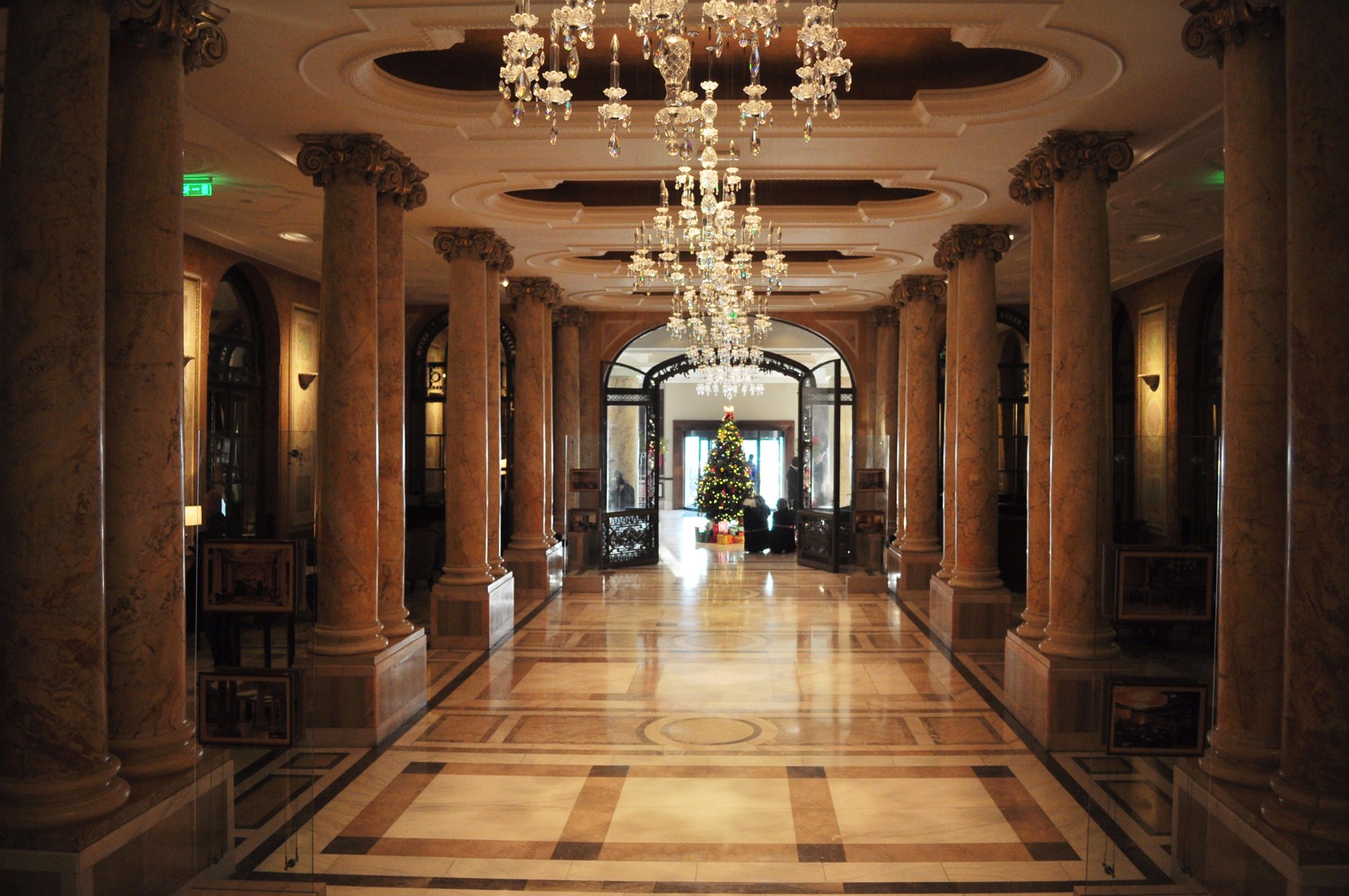 A wide view through the grand Athénée Palace hotel lobby, with marble columns, chandeliers, and the entrance beyond
