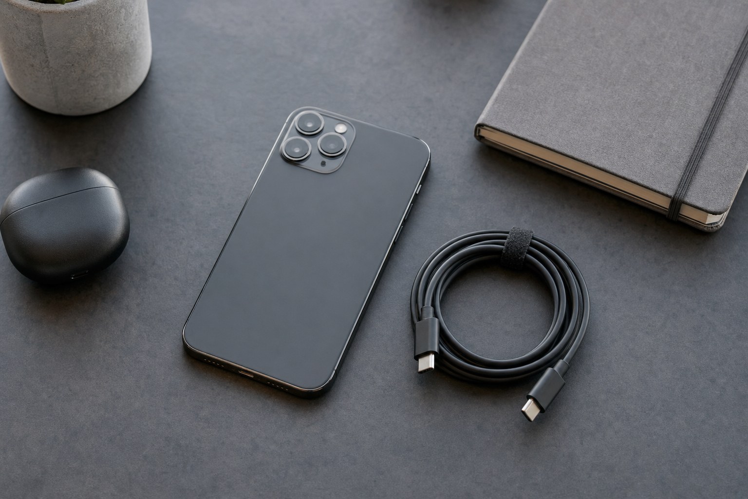 Phone, USB-C cable, earbud case, and notebook on a dark desk