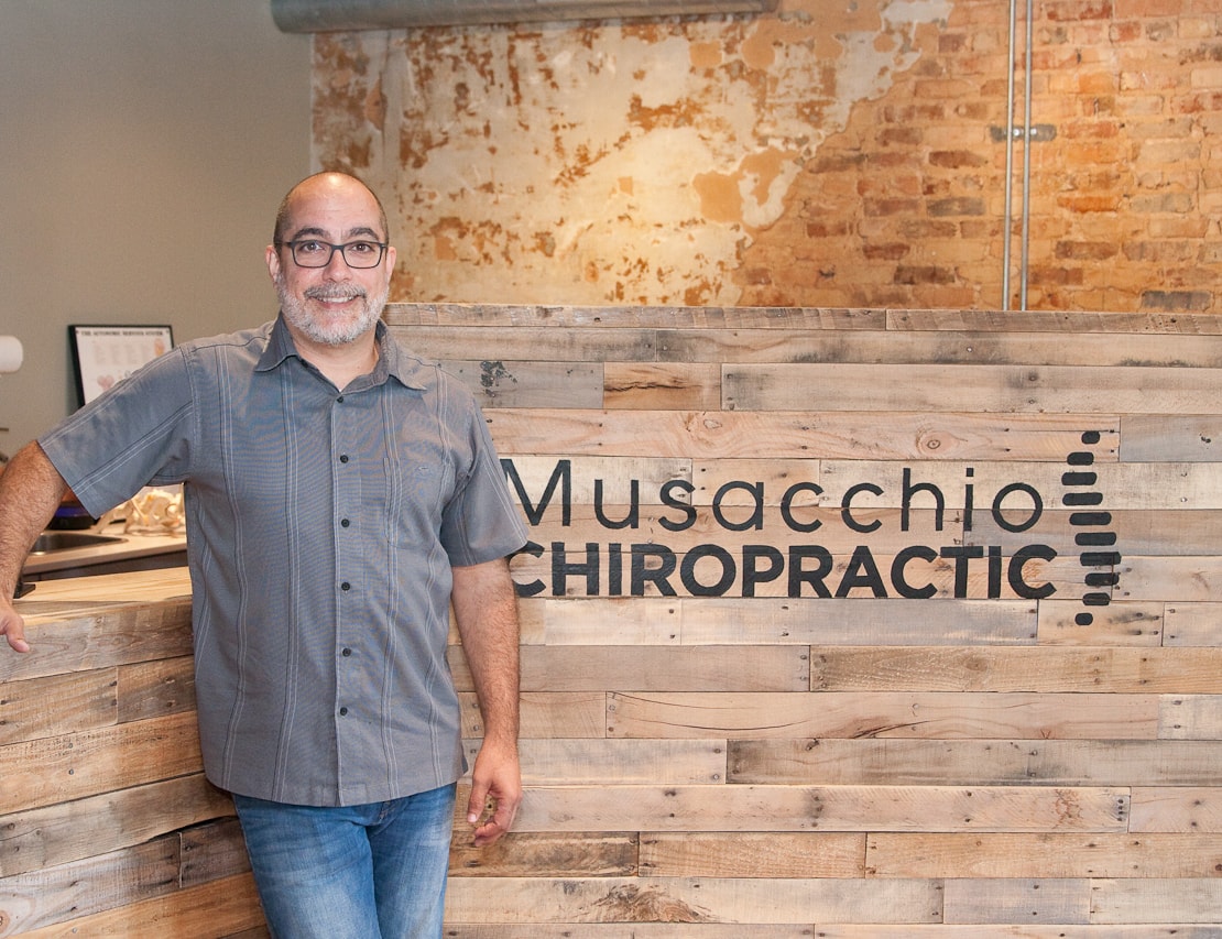 Dr. Chris Musacchio standing in front of the Musacchio Chiropractic logo wall